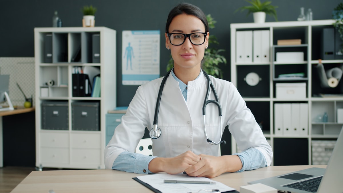 Médica sorridente, de jaleco branco e estetoscópio, em consultório